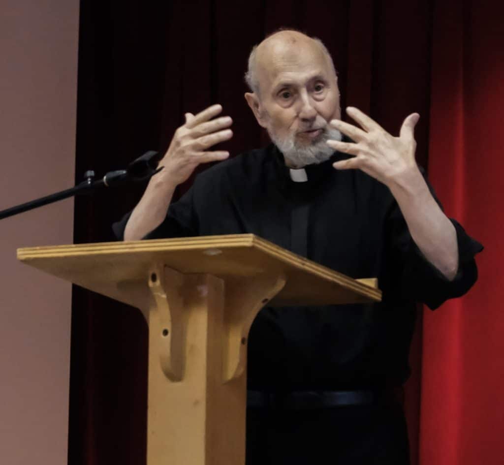 Father George Makhlouf, dressed in a black clergy outfit, stands at a podium speaking passionately to a crowd.