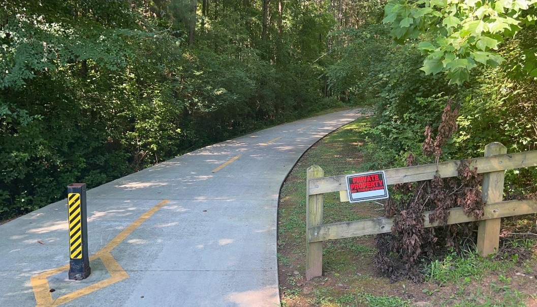 A small, generic sign reading "PRIVATE PROPERTY - NO TRESPASSING" has been affixed to a fence near a pedestrian walkway leading to Intrenchment Creek Park in DeKalb County, GA.