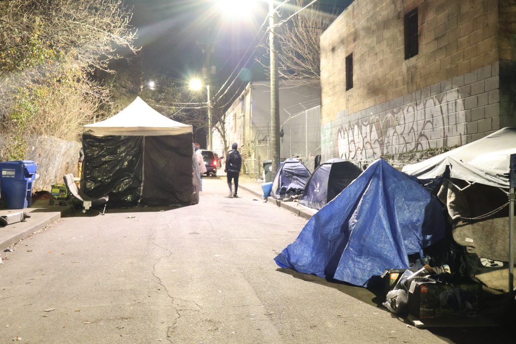 Image of a street at night, taken from the middle of the road. The street is lit by a bright streetlamp at the top center of the image. On the right-hand side are tents used by unhoused Atlantans as shelter. Further up the street on the left is a temporary warming shelter created by activists. An individual in dark clothing is walking in the street at the center of the image. 