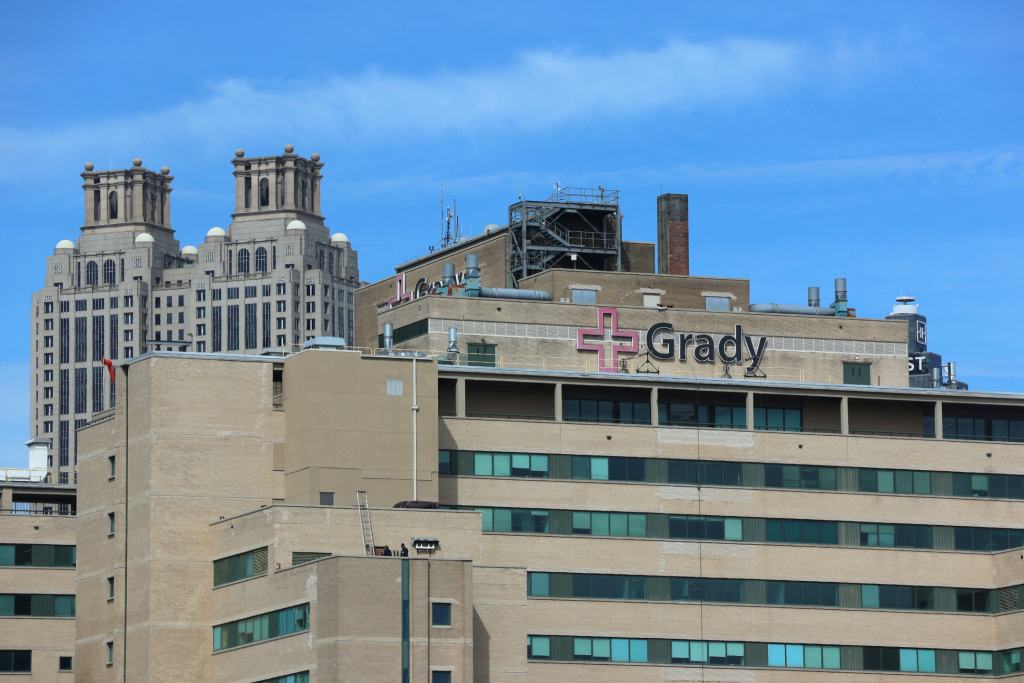 The upper half of Grady Hospital where Doctors are combatting climate change health risks in Downtown Atlanta. The hospital facade is brown brick and concrete. At the top of the building sits signage that includes the international symbol for hospitals with "Grady" to its right from the picture's perspective. It is a clear day with blue skies and few wispy clouds. In the background sits one of Atlanta's shorter skyscrapers with art-deco architecture.