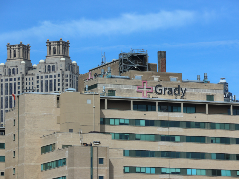 The upper half of Grady Hospital where Doctors are combatting climate change health risks in Downtown Atlanta. The hospital facade is brown brick and concrete. At the top of the building sits signage that includes the international symbol for hospitals with "Grady" to its right from the picture's perspective. It is a clear day with blue skies and few wispy clouds. In the background sits one of Atlanta's shorter skyscrapers with art-deco architecture.