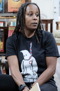 Erin Parks, a Black woman, wears a black shirt with a hooded-Ku Klux Klan figure hanged by a rope. Parks sits in front of bookshop shelves. 