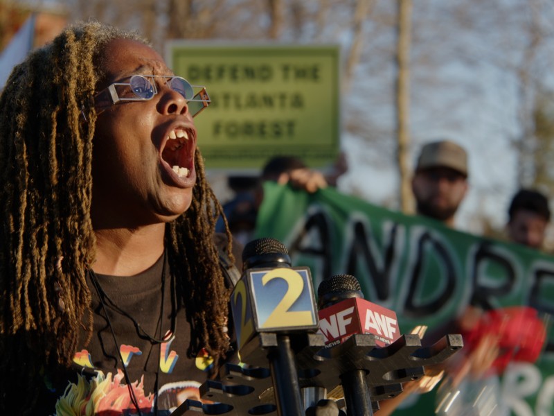 Dawn O’Neal of the People’s Campaign to Stop Cop City speaks passionately into a Channel 2 microphone while a group of people who oppose Andre Dickens support her in the background.