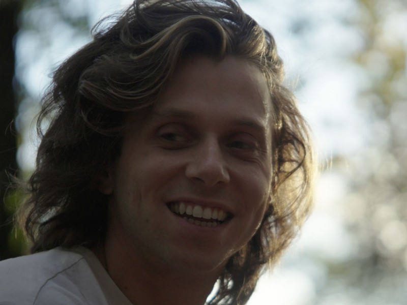A cinematic still of Jamie Mariscano, a white nonbinary person with medium-length dark blonde hair wearing a white t-shirt. Jamie is smiling. The setting is close to dusk, with long shadows. The sun frames left side of Jamie's face. Only the upper quarter of Jamie is visible. The setting is woodsy, with trees visible but slightly blurry in the background. The