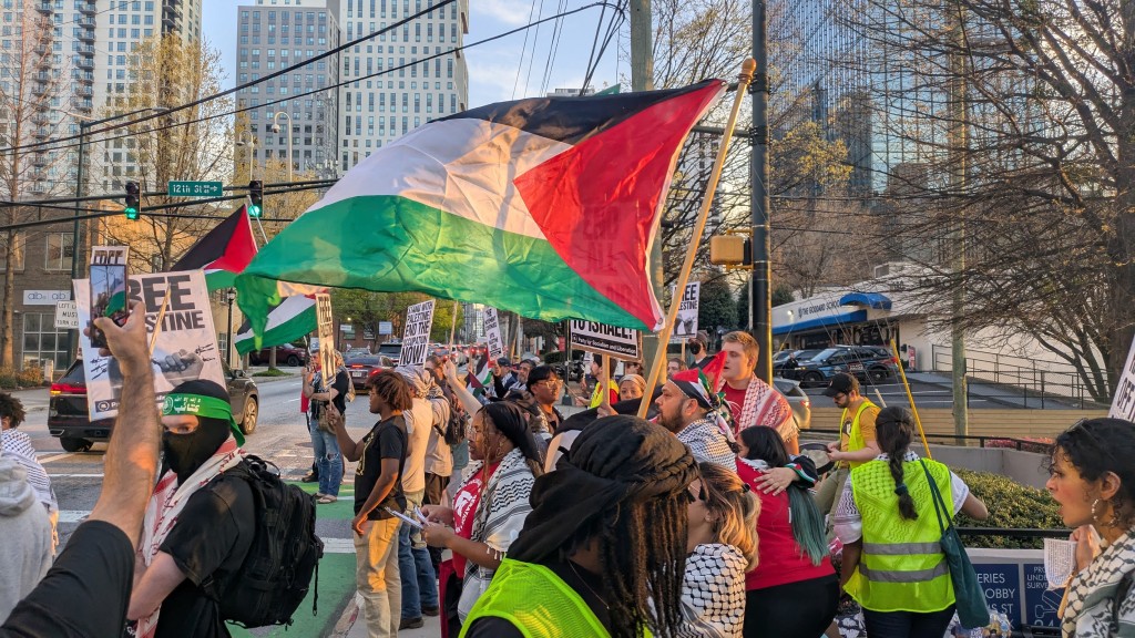 Protesters gathered in a city street holding Palestinian flags and signs reading 'Free Palestine' during a demonstration. Skyscrapers and city buildings are visible in the background under a clear sky. Many protesters are wearing Kefiyehs, some are dressed in neon safety vests and one is dressed as a Hamas fighter with a black balaclava and green bandana.