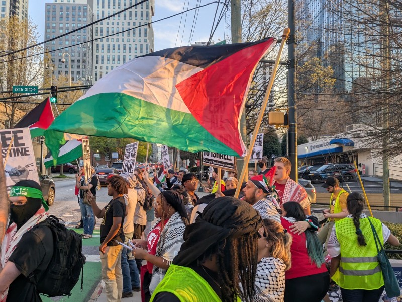 Protesters gathered in a city street holding Palestinian flags and signs reading 'Free Palestine' during a demonstration. Skyscrapers and city buildings are visible in the background under a clear sky. Many protesters are wearing Kefiyehs, some are dressed in neon safety vests and one is dressed as a Hamas fighter with a black balaclava and green bandana.