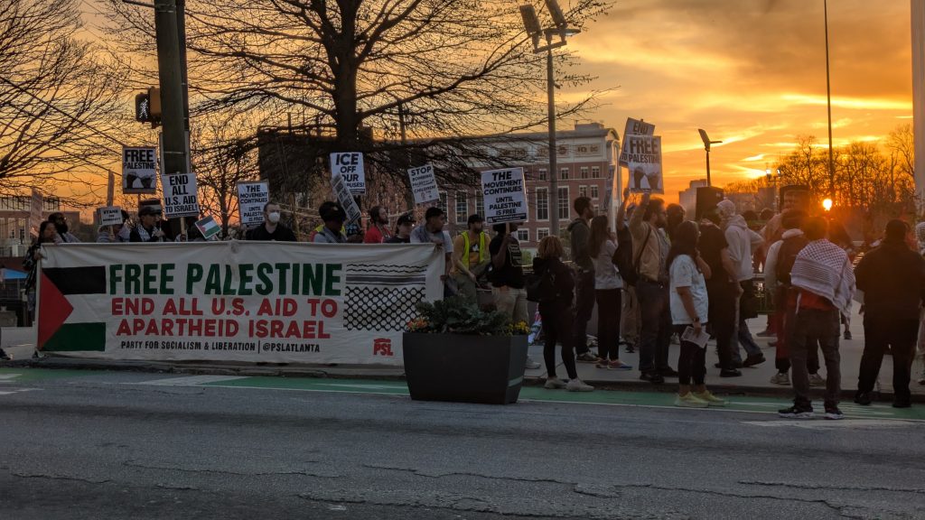 Protesters holding signs and a large banner reading 'Free Palestine - End All U.S. Aid to Apartheid Israel' during a demonstration outside the Israeli consulate in Midtown Atlanta at sunset.