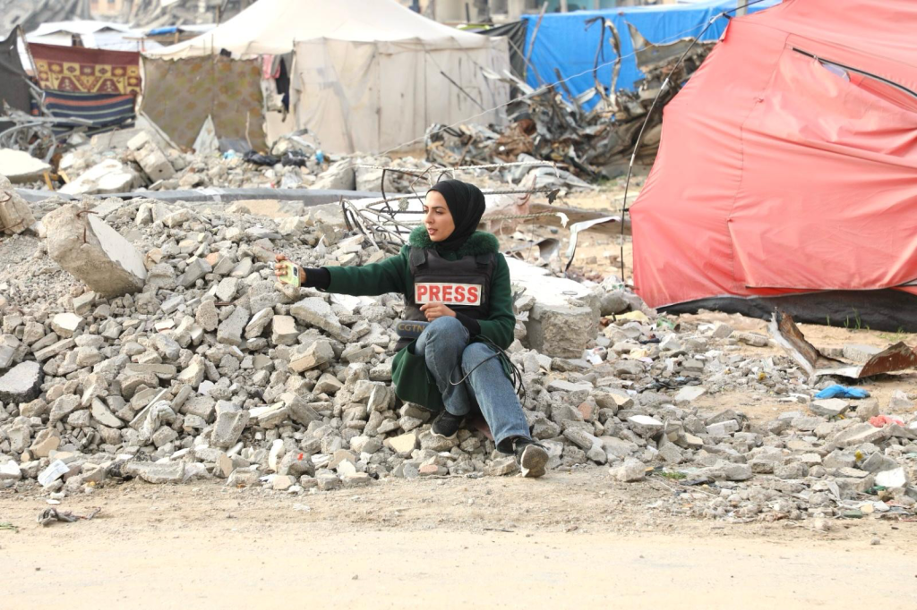Female journalist wearing a press vest and headscarf sits on rubble, holding a phone, surrounded by tents and debris in Gaza.