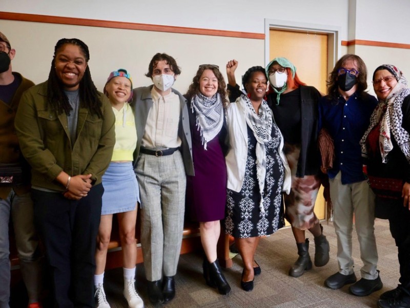 A group of nine smiling people wearing business casual attire stands in a curved line inside a beige hallway. The group is of mixed genders and races. The three individuals in the middle are the "Weealune Three" Cop City protesters. Left to right they are Noah Grigni wearing grey slacks, a pink oxford, grey jacket and KN-95 mask; Krya Hanon, wearing a purple dress and a keffiyeh, and Rukia Rogers, wearing a patterned black dress and keffiyeh.