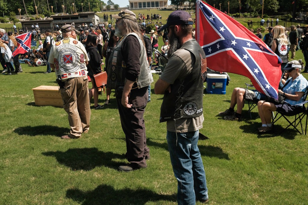 Dozens of participants, some of whom are wearing motorcyle club vests—including one for the Sons of Confederate Veterans Mechanized Calvary—stand near a Confederate flag at the 2022 Confederate Memorial Day rally in Stone Mountain, Georgia.