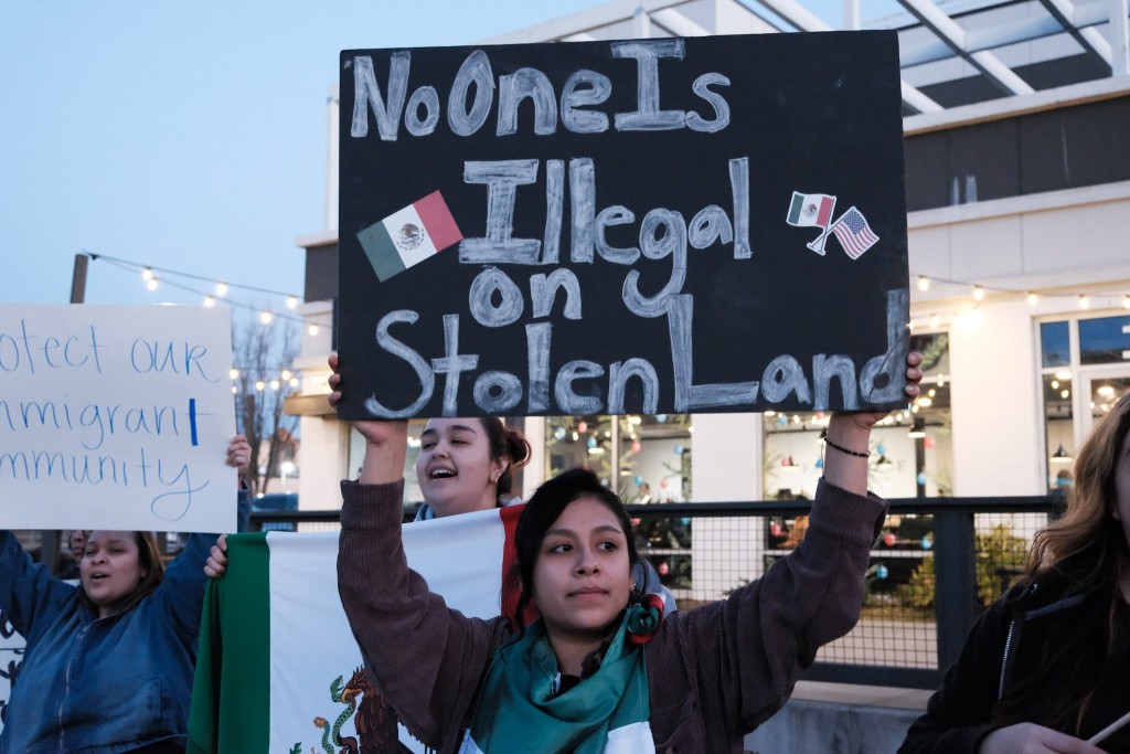 A woman stands at a rally holding a sign that says, "No one is illegal on stolen land."