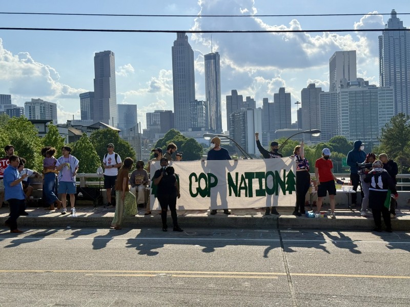 Protesters on Jackson Street Bridge hold a "No Cop Nation Banner" with Atlanta's skyline visible in the background.